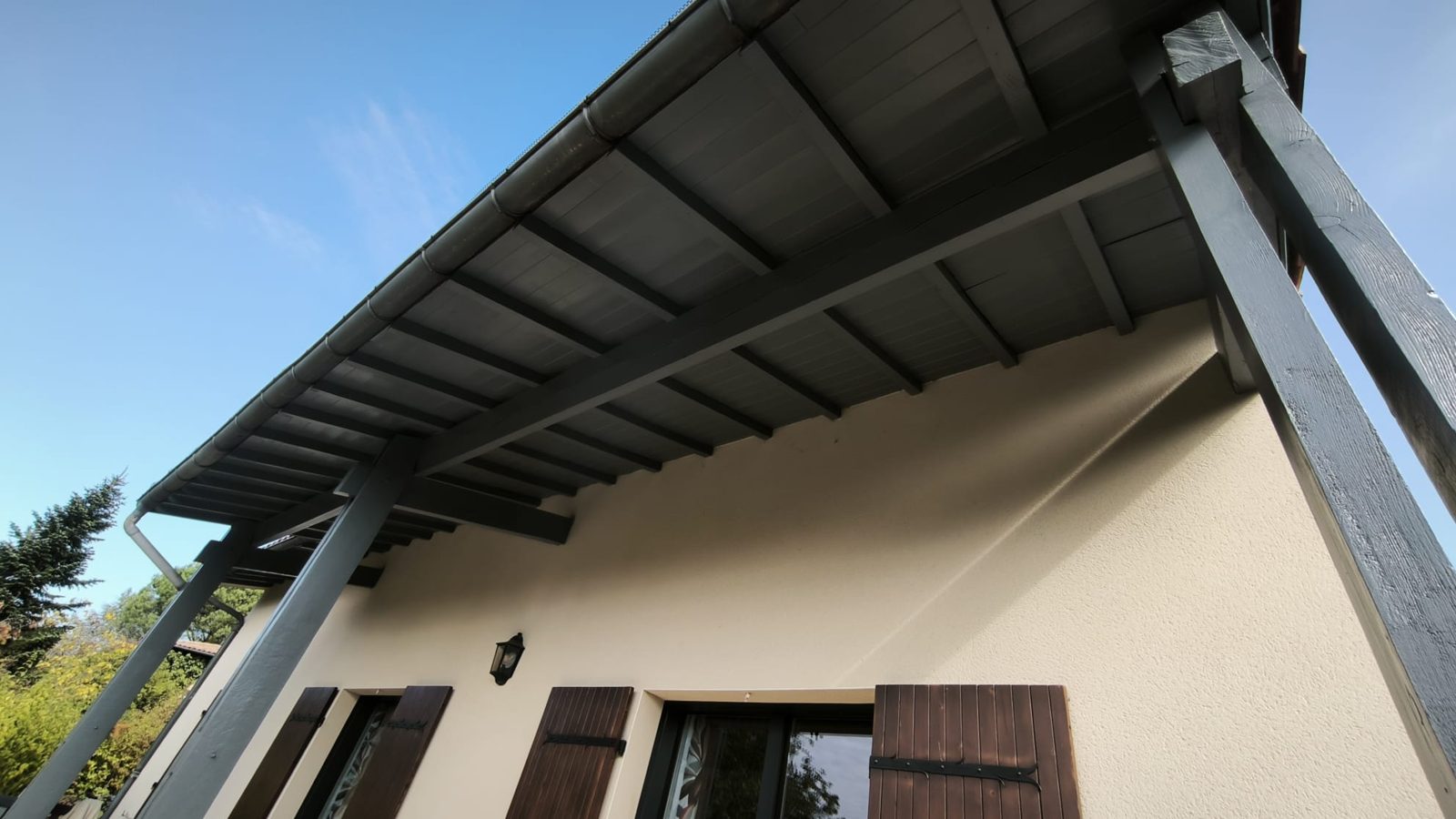 Avancée de toit et poteaux en bois repeints en gris, au-dessus d'une façade claire aux volets bruns